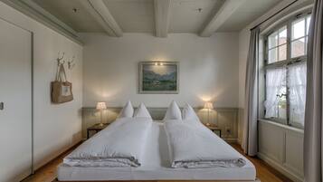 A double bed with white linens, two bedside tables with lamps, a painting on the wall, and a window with curtains.
