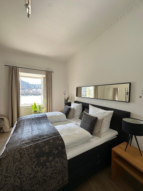 A hotel room with a large bed, a bedside table, a mirror, and a window with curtains. - Hotel Schloss Ort (Passau)