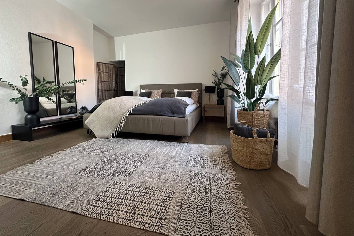 A modern bedroom with a large bed, a bedside table, a mirror, and a potted plant.