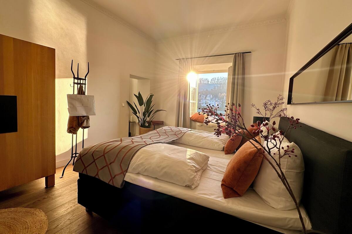 A bedroom with a bed, a bedside table, a mirror, and a window with curtains.