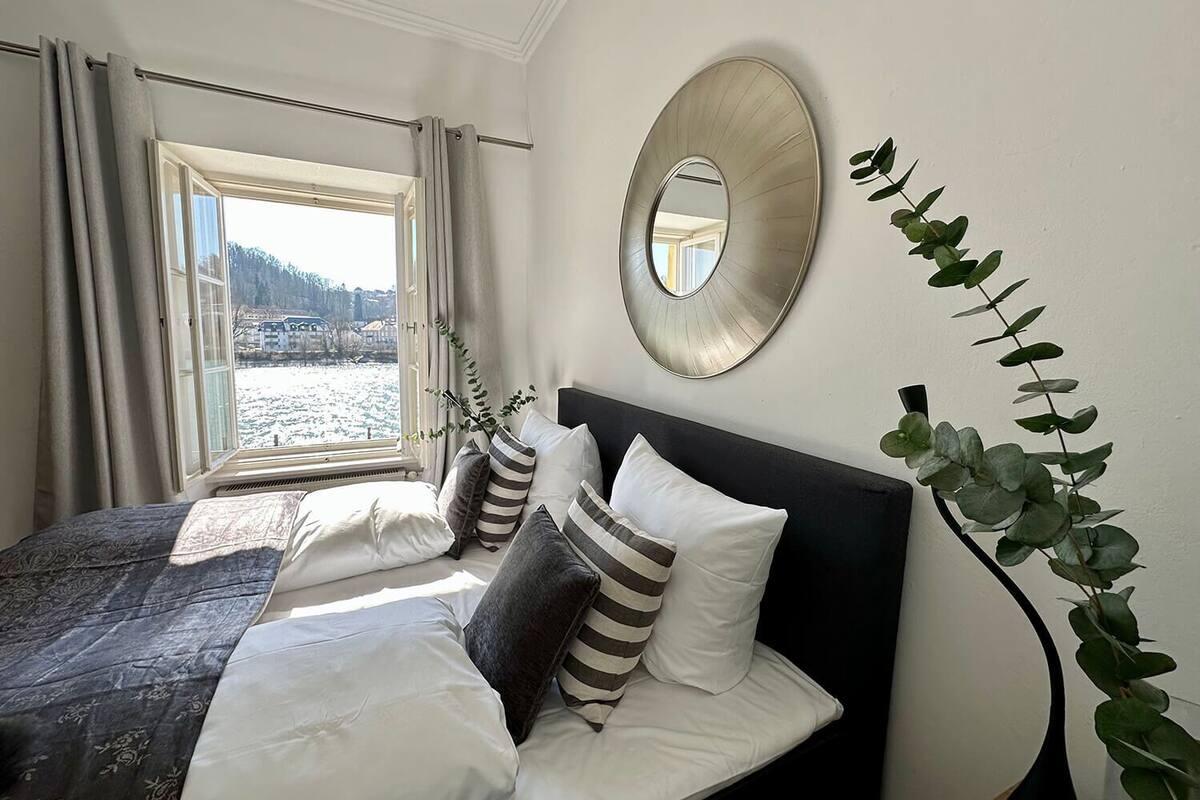 A bedroom with a bed, a window with curtains, a mirror, and a plant.