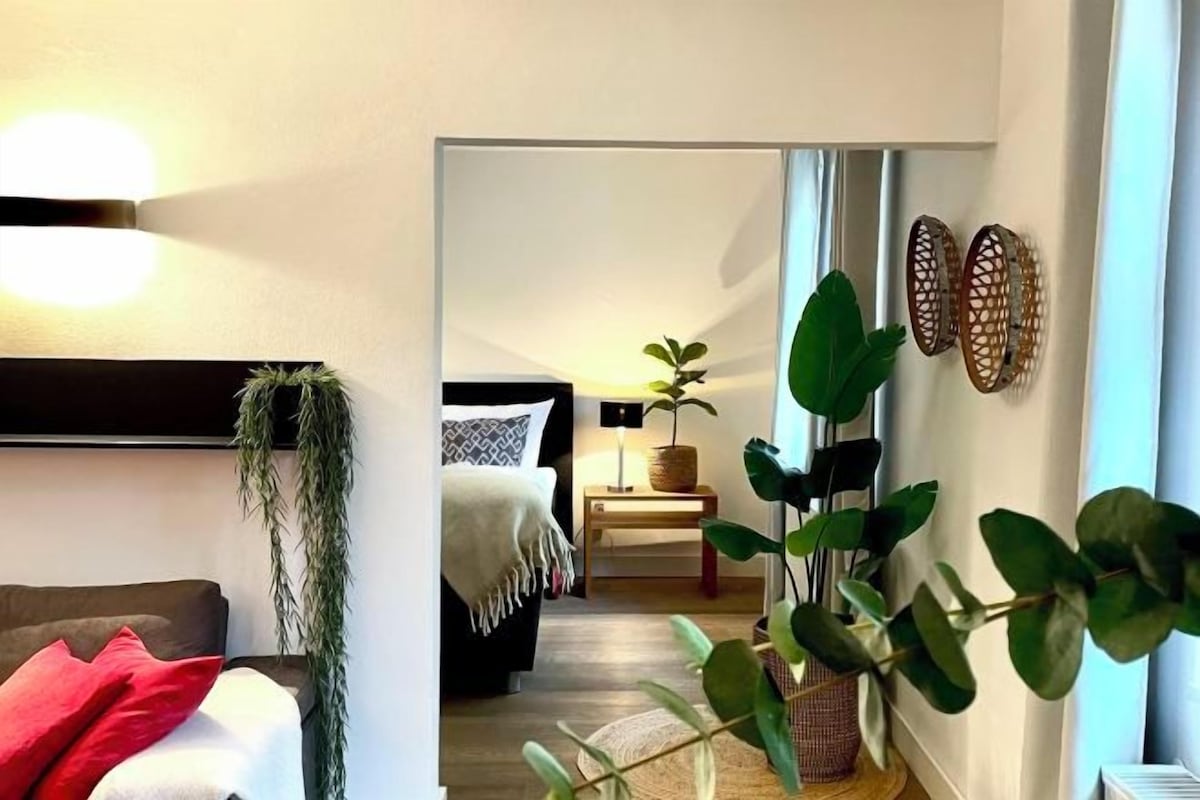 A modern living room with a sofa, a coffee table, and a plant. There is a bed with a nightstand and a lamp in the background.