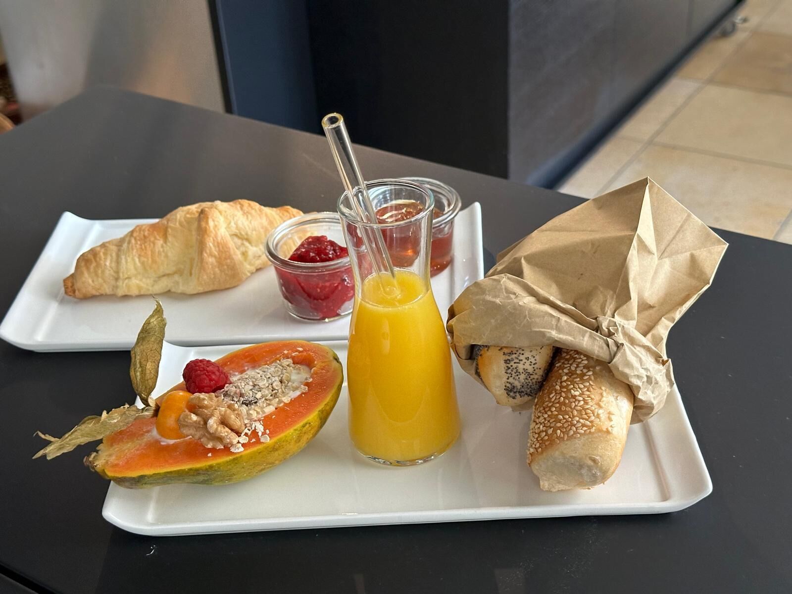 Un plato de desayuno con un croissant, media papaya con semillas, un vaso de zumo de naranja y un sándwich envuelto en papel.