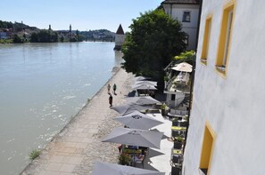 A riverside walkway with outdoor seating and umbrellas, overlooking a river with buildings and a tower in the background. - Hotel Schloss Ort (Passau)