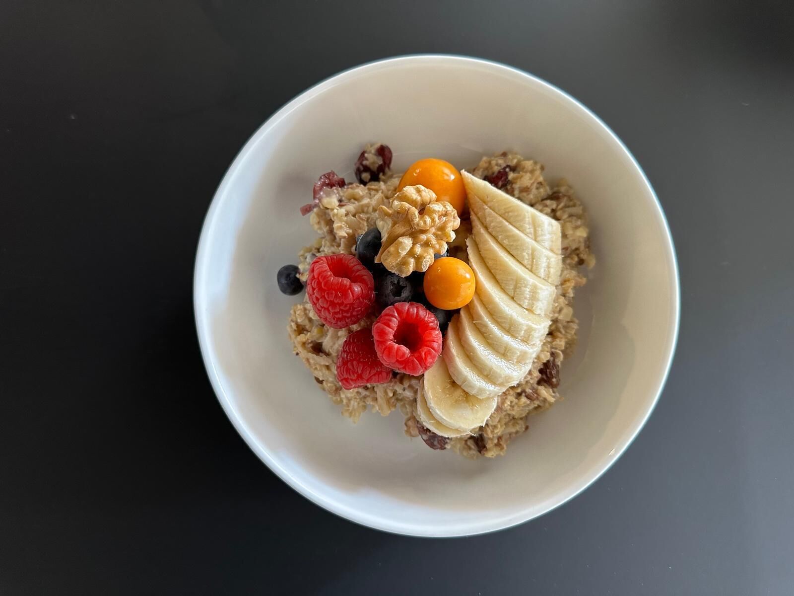 Un bol de porridge garni de tranches de banane, de framboises, de myrtilles et d’une noix, le tout posé sur une surface sombre.