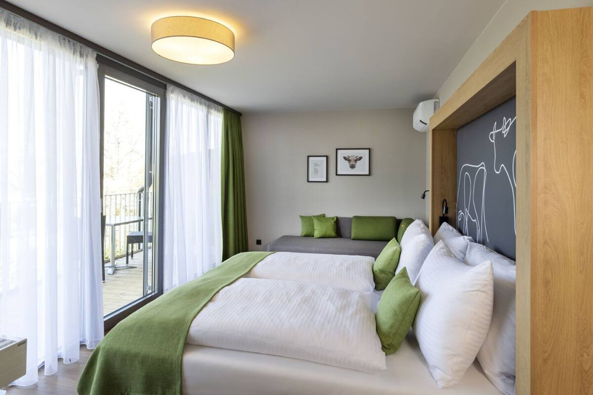 A modern bedroom with a large bed, green and white bedding, a grey sofa, and a view of a balcony.
