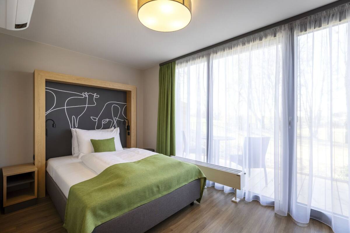 A modern hotel room with a large bed, a headboard with a unique design, a bedside table, and a view of the outdoors through sheer curtains.
