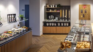 Zona de bufé con diversos alimentos, entre ellos pasteles y pan, y una estación de café con múltiples dispensadores.