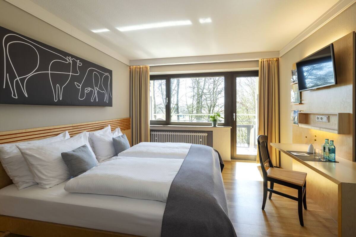 A hotel room with a large bed, a desk, a TV, and a window with a view of trees.