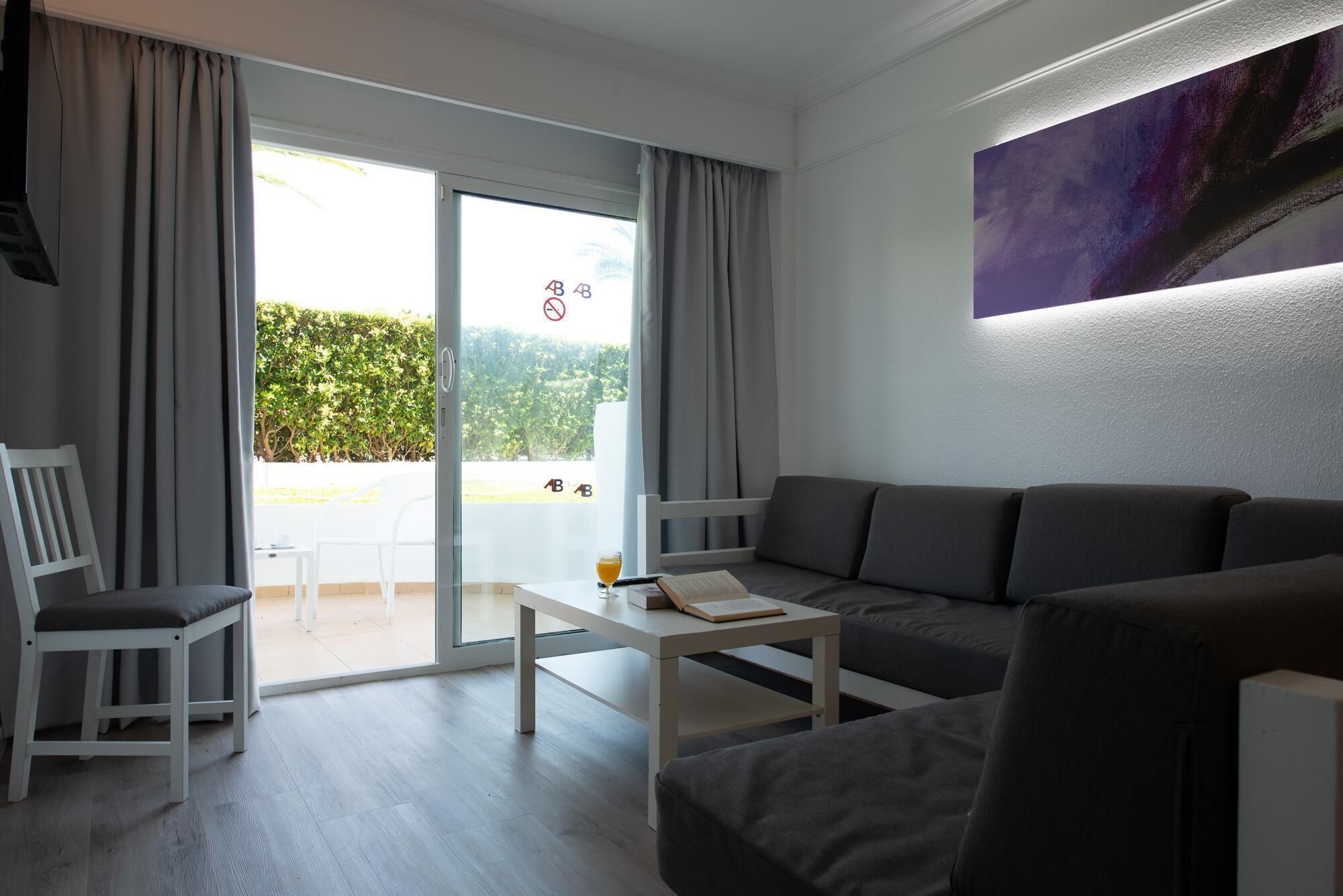 A modern living room with a sofa, a small table, and a chair. There is a sliding glass door leading to an outdoor area with a view of greenery.