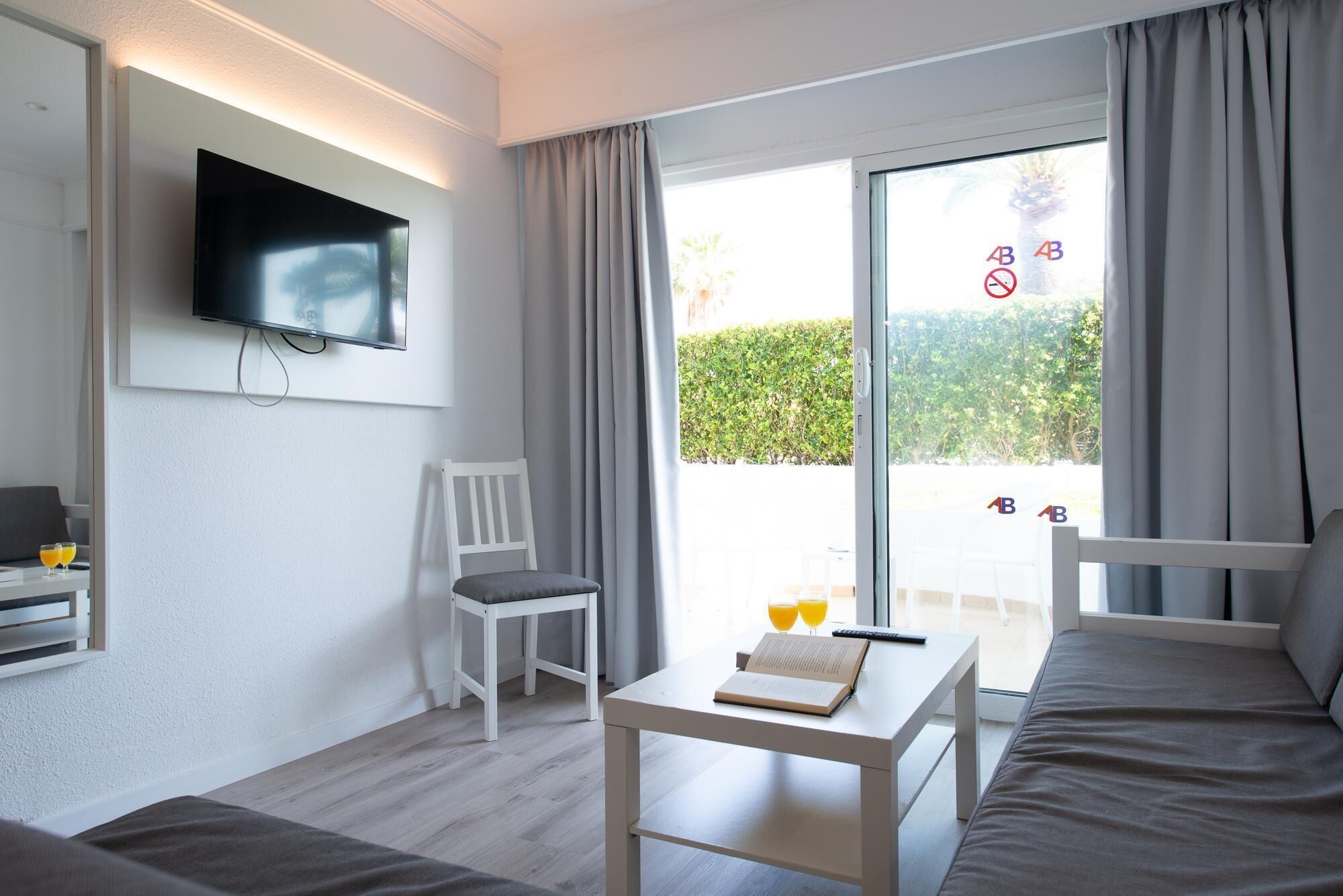 Una habitación de hotel moderna con un televisor de pantalla plana montado en la pared, una silla blanca, una mesita con un libro y gafas, y una cama.