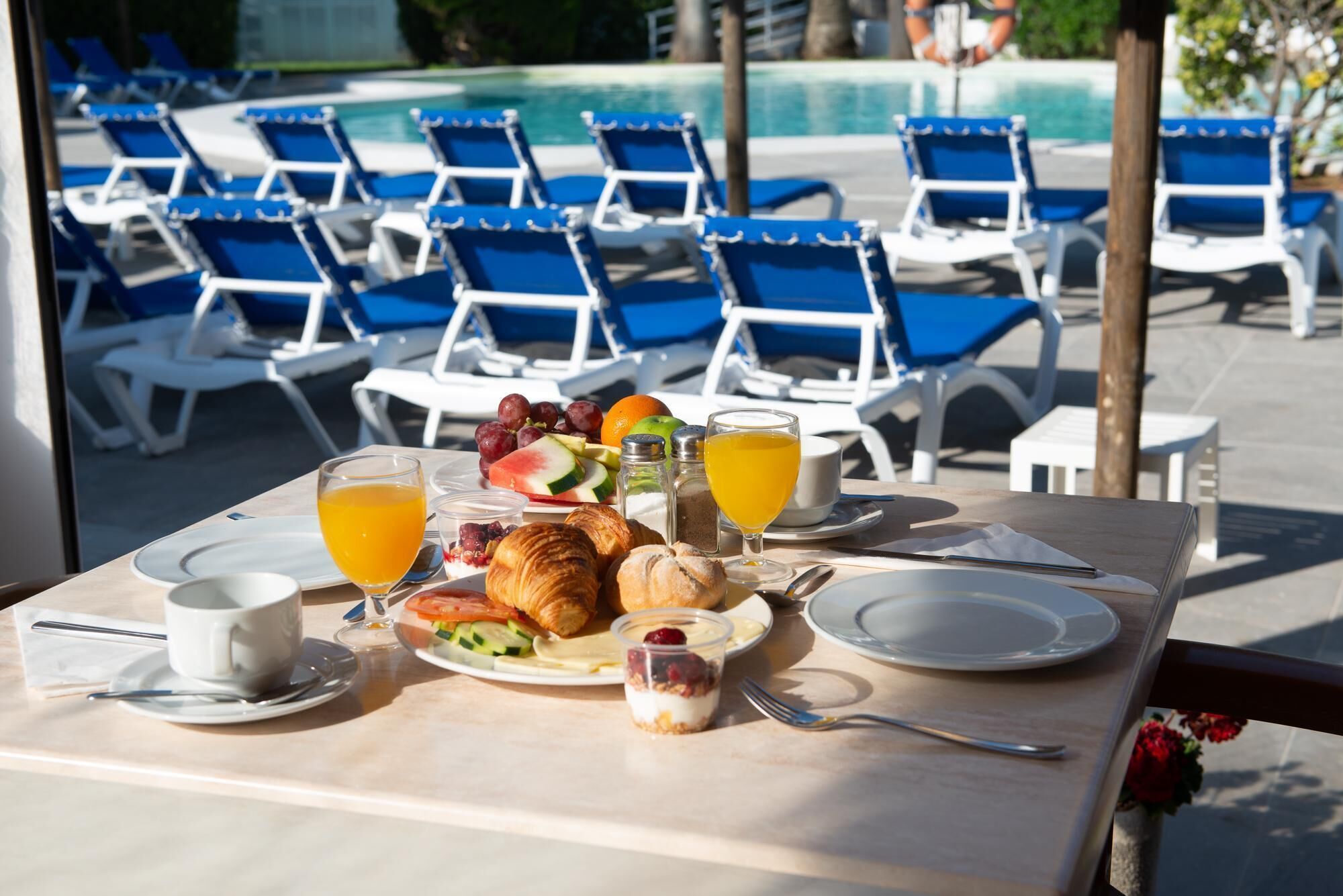 A breakfast table with croissants, fruit, and juice, overlooking a pool with blue chairs.