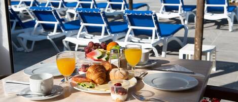 Een ontbijttafel met croissants, fruit en sap, met uitzicht op een zwembad met blauwe stoelen.