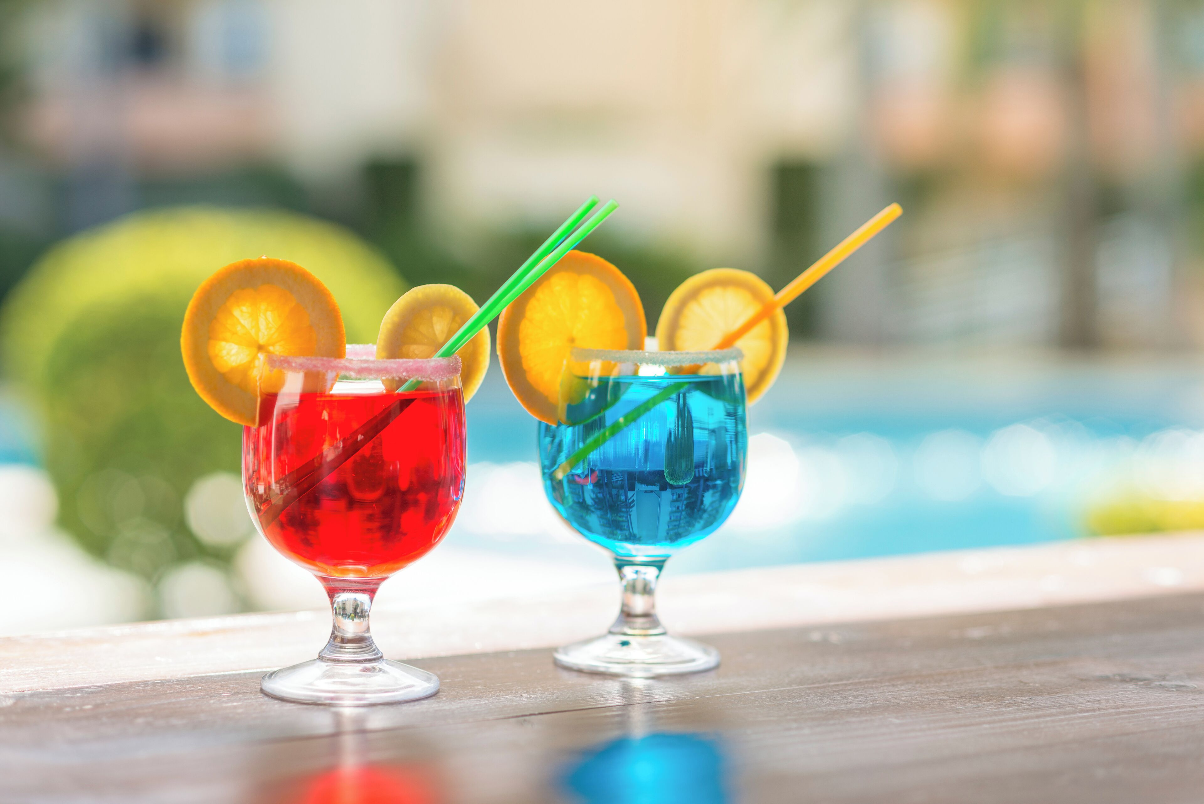 Zwei Cocktails mit Obstdekoration auf einer Holztischplatte in der Nähe eines Pools.