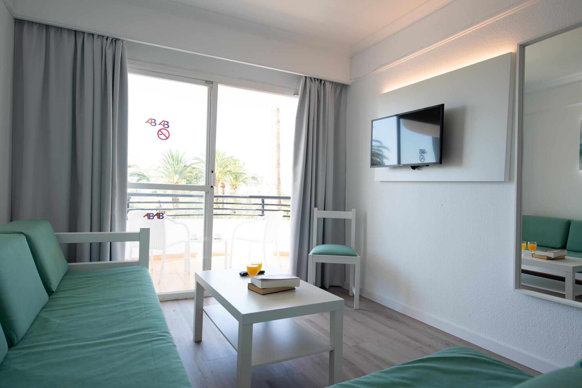 A modern hotel room with a balcony, a TV, a sofa, a table with a book and a glass of orange juice, and a green cushion.