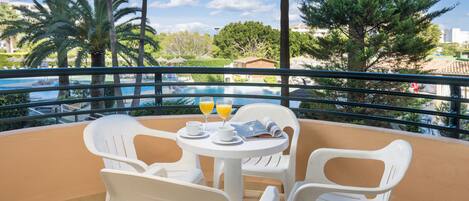A balcony with a table, chairs, and a view of a swimming pool and palm trees.