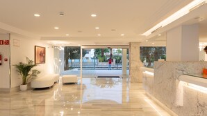 Un moderno vestíbulo de hotel con un mostrador de recepción de mármol, un área de descanso con sofá y una planta en maceta, y un puerta de cristal que da al exterior.