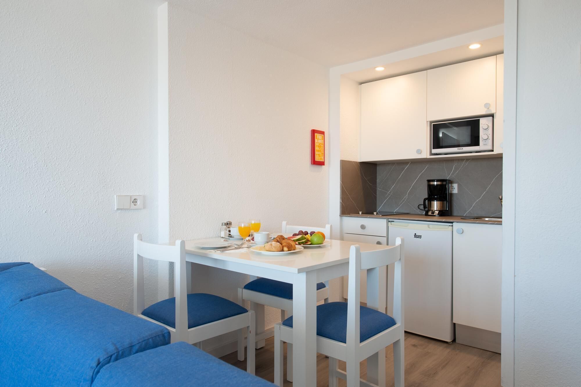 Une petite cuisine équipée d’un four à micro-ondes, d’une machine à café et d’un petit réfrigérateur. On y trouve une table à manger avec des chaises, ainsi qu’un canapé bleu dans l’espace salon.
