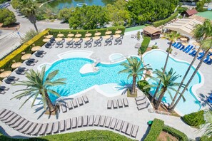 Eine Vogelperspektive zeigt ein Resort mit einem großen Schwimmbad, Liegestühlen, Palmen und einer gepflegten Landschaft.