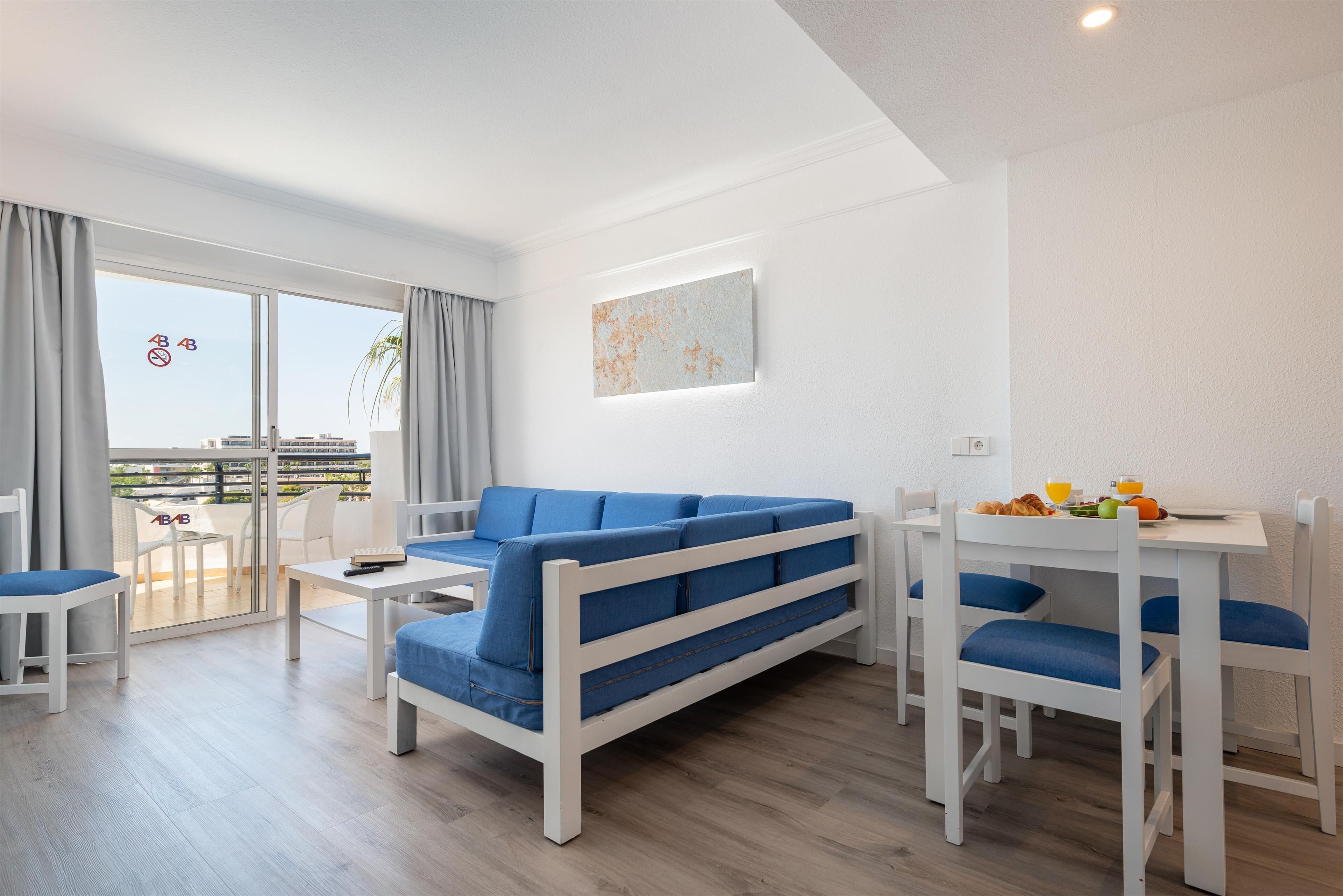 A modern living room with a blue sofa, white dining table, and chairs. There is a glass door leading to a balcony with a view of buildings.