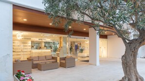 Un edificio moderno con fachada de vidrio, zona de estar al aire libre con mobiliario de mimbre y un árbol en primer plano.