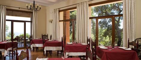A dining area with tables set for a meal, large windows with curtains, and a chandelier.
