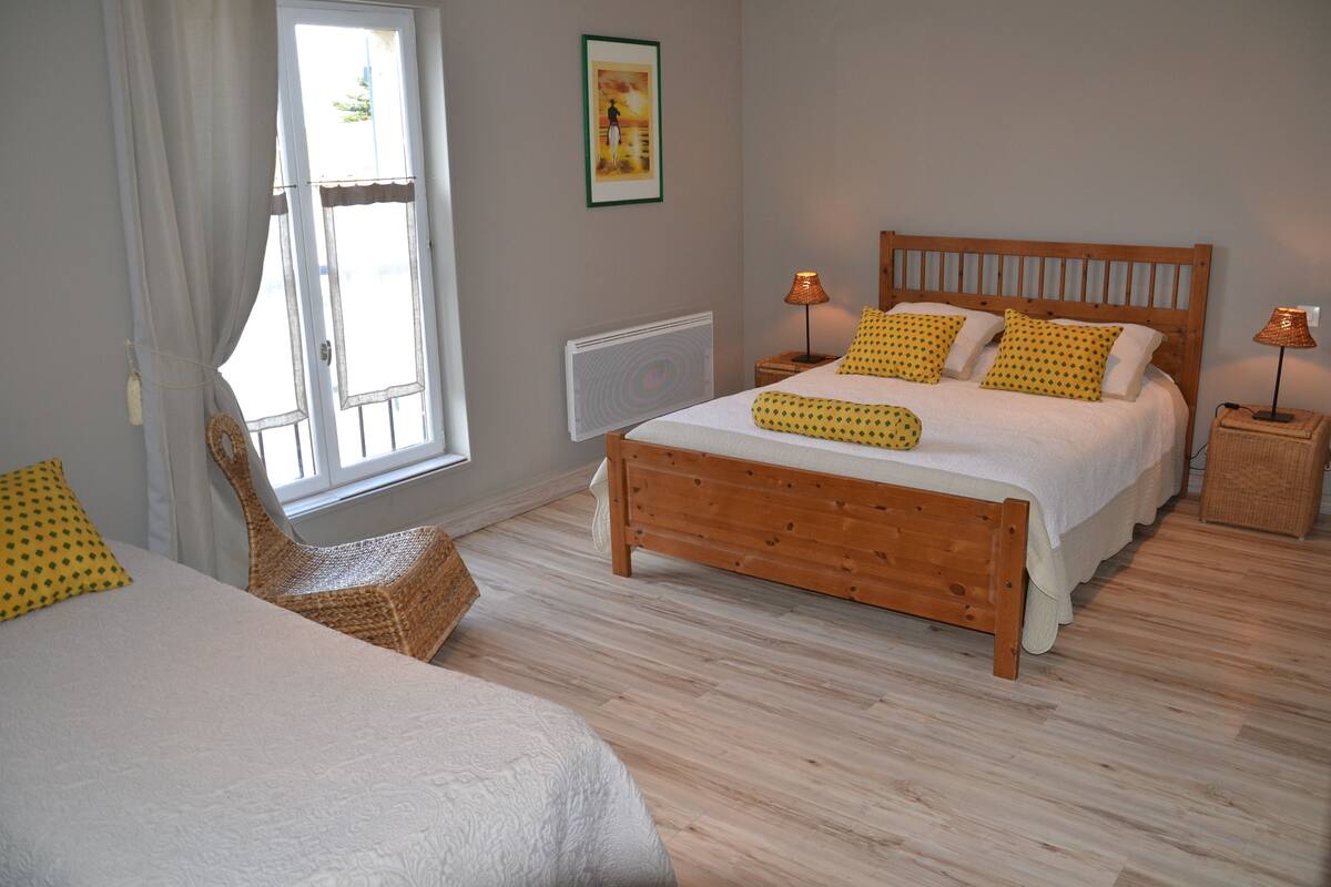 Un dormitorio con una cama de madera, dos mesitas de noche con lámparas, una silla de mimbre y una ventana con cortinas.