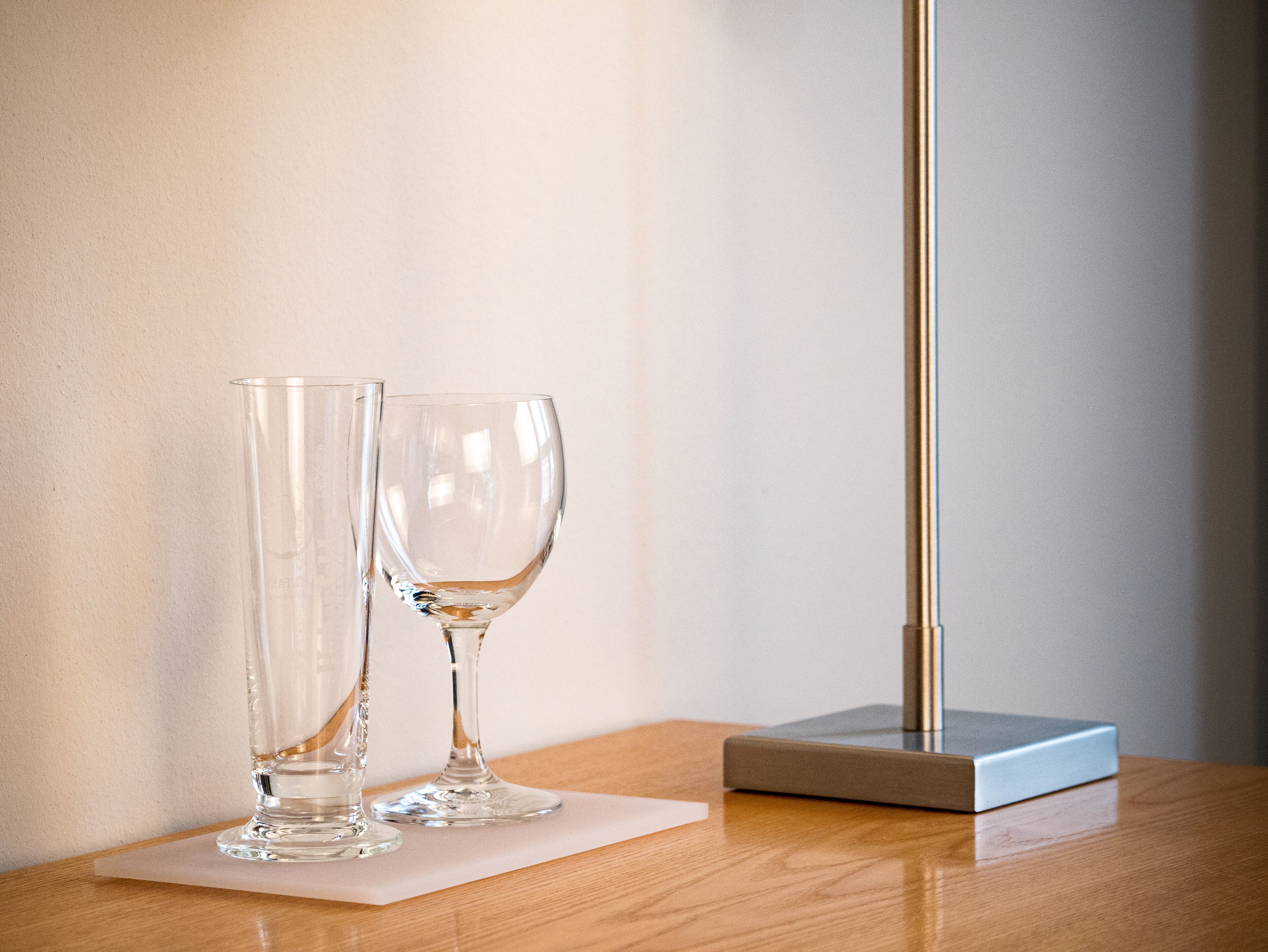 Zwei klare Gläser auf einem Holztisch, mit einer weißen Serviette, einem silbernen Gegenstand und einer hohen Lampe vor einer schlichten Wand.