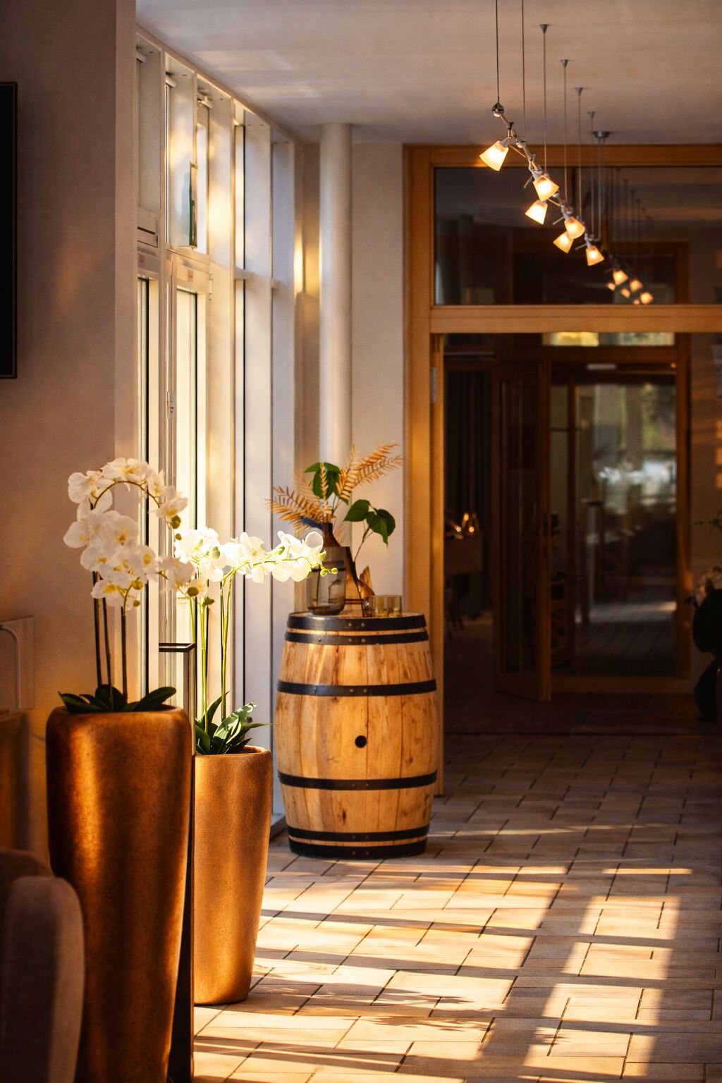 A hallway with large windows, a wooden barrel, and potted orchids.