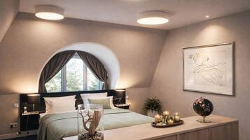 A bedroom with a large bed, a bedside table, a desk, a window with curtains, and a framed artwork on the wall.