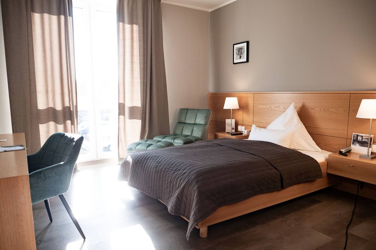 A hotel room with a bed, a desk, a chair, and a nightstand with a lamp.