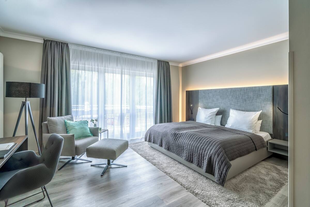 A modern hotel room with a large bed, a desk, a chair, and a view of the city through large windows.