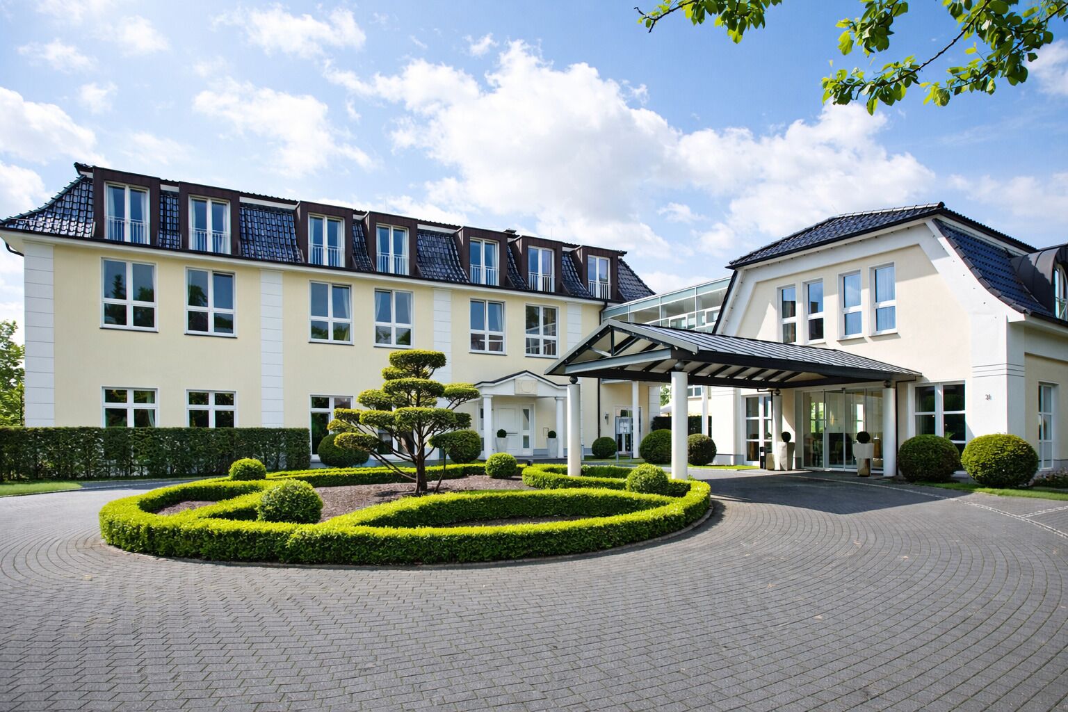 A hotel with a symmetrical facade, a covered entrance, and a landscaped garden with a central tree.