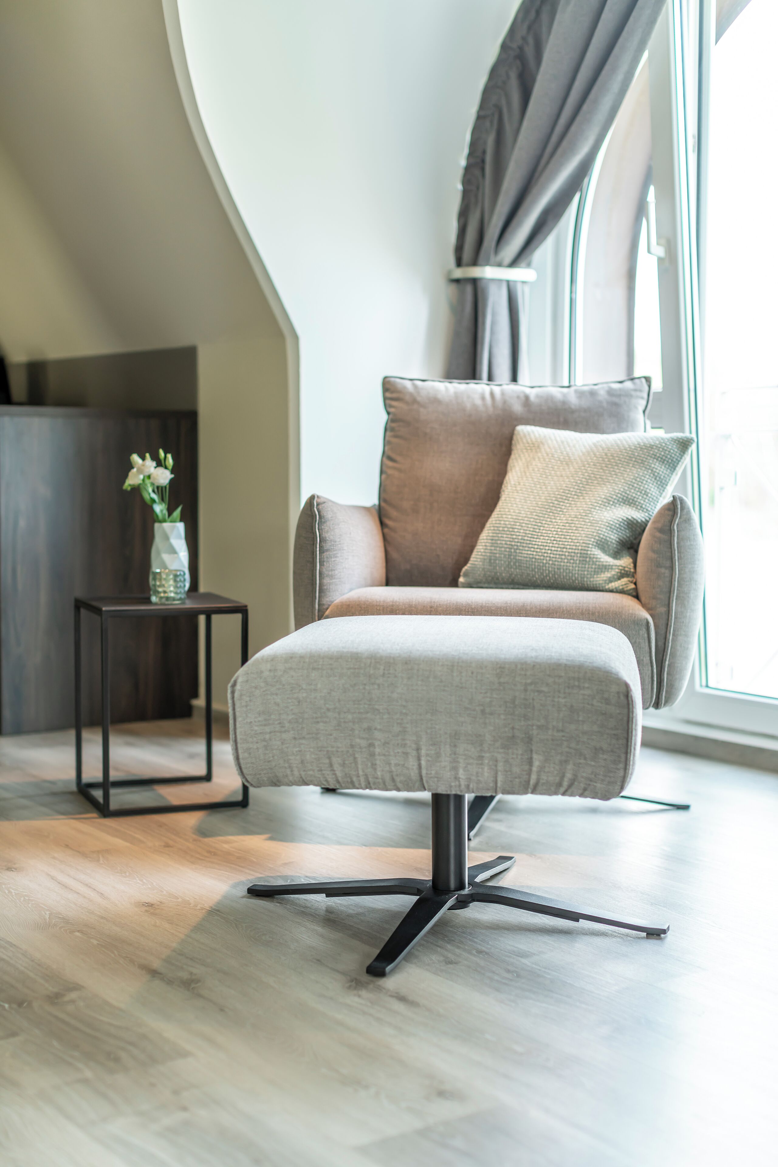 Un sillón moderno con base metálica, ubicado en una habitación con suelo de madera y una mesita auxiliar con un florero.