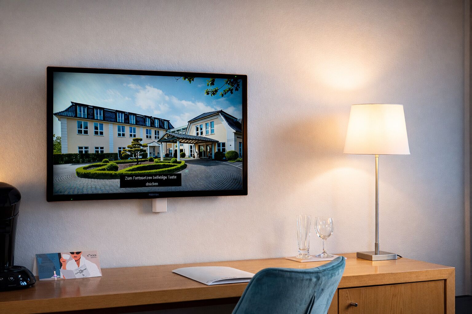 Ein Hotelzimmer mit Fernseher, der ein Gebäude zeigt, einem Schreibtisch mit Lampe und einem Sessel.