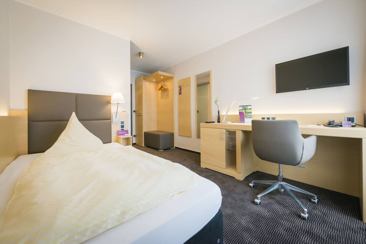 A hotel room with a large bed, a desk with a chair, a flat-screen TV, and a small bathroom.