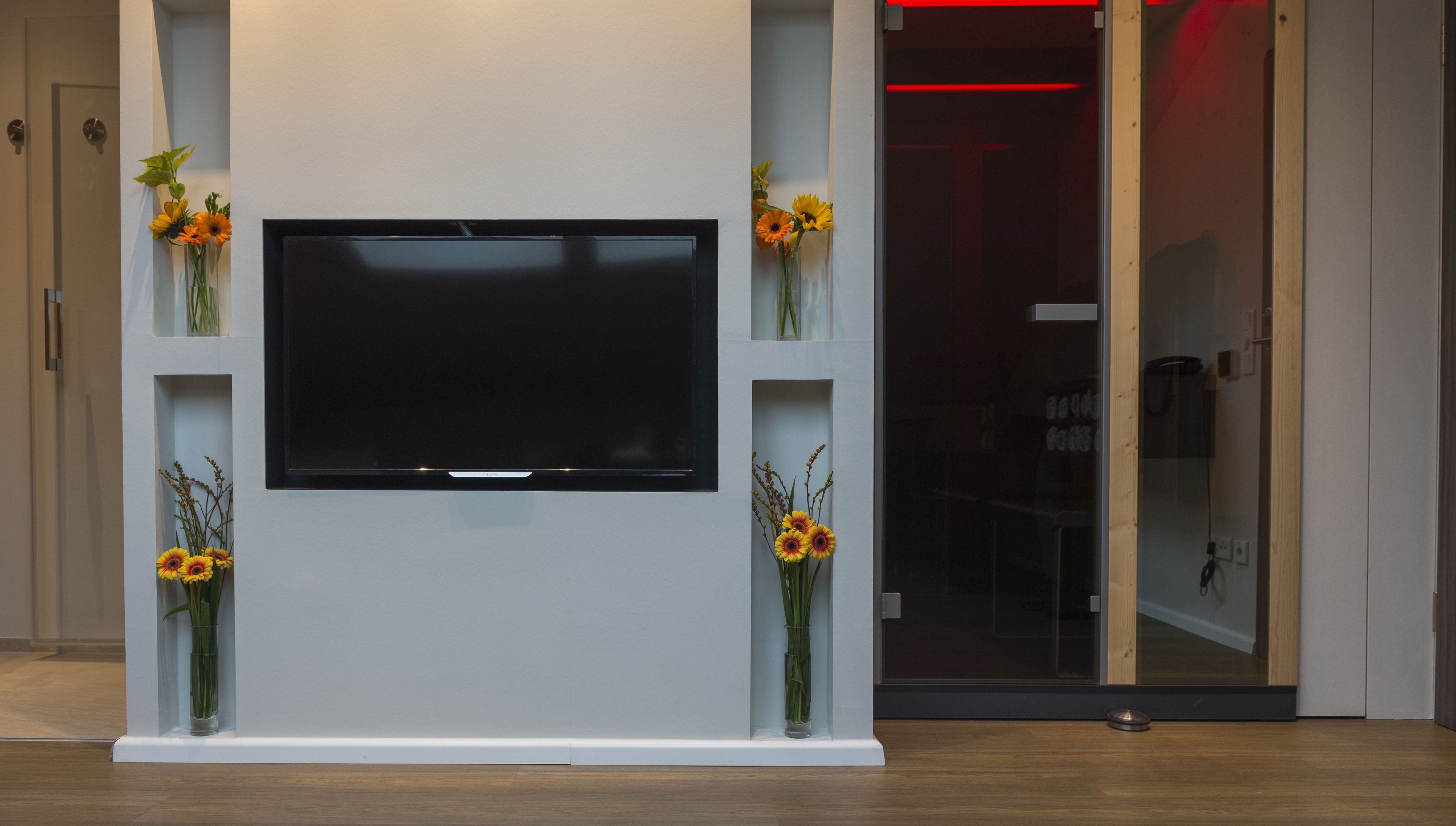 Ett modernt vardagsrum med en platt-TV som är monterad på en vit vägg, och som omges av två hyllor i glas, där blomsterarrangemang är placerade.