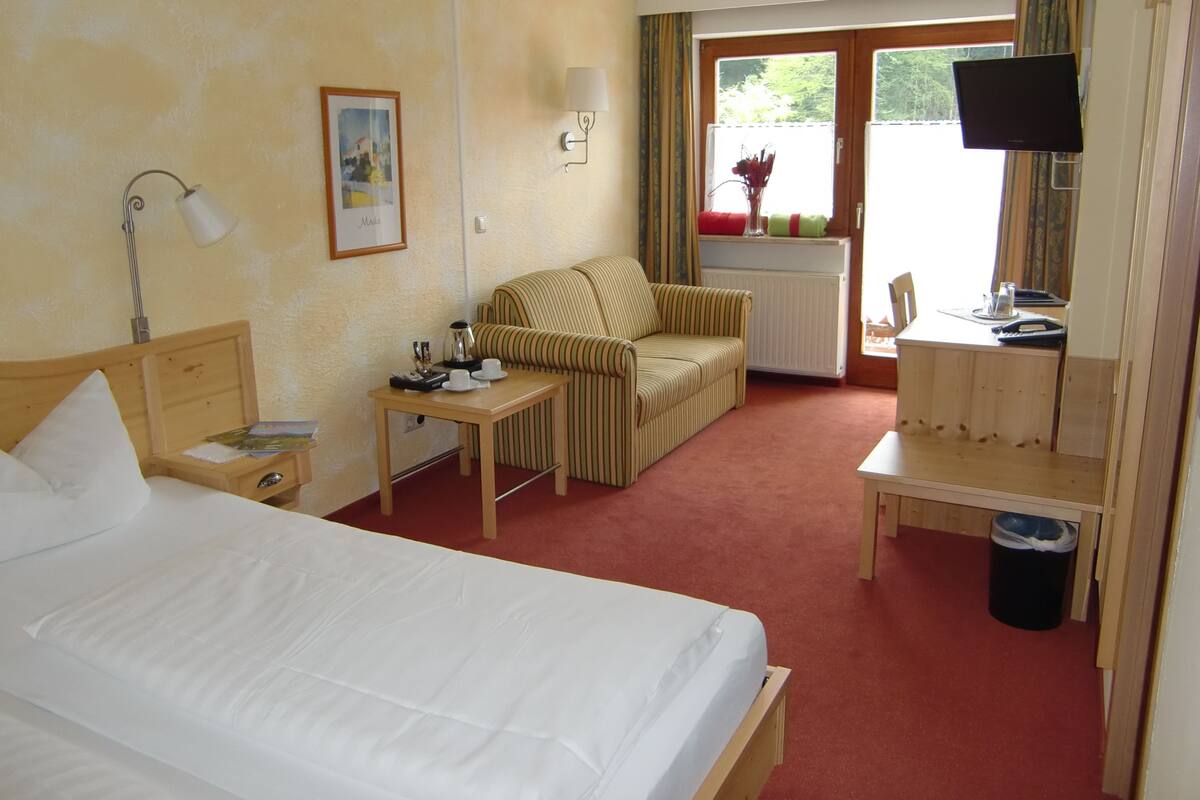 Ein Hotelzimmer mit einem Bett, einem kleinen Tisch mit Lampe, einem gestreiften Sessel, einem Fernseher und einem Fenster mit Vorhängen.