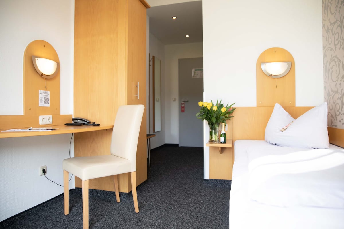 Quarto de hotel com escrivaninha, cadeira, cama e mesa de cabeceira com um vaso de flores.