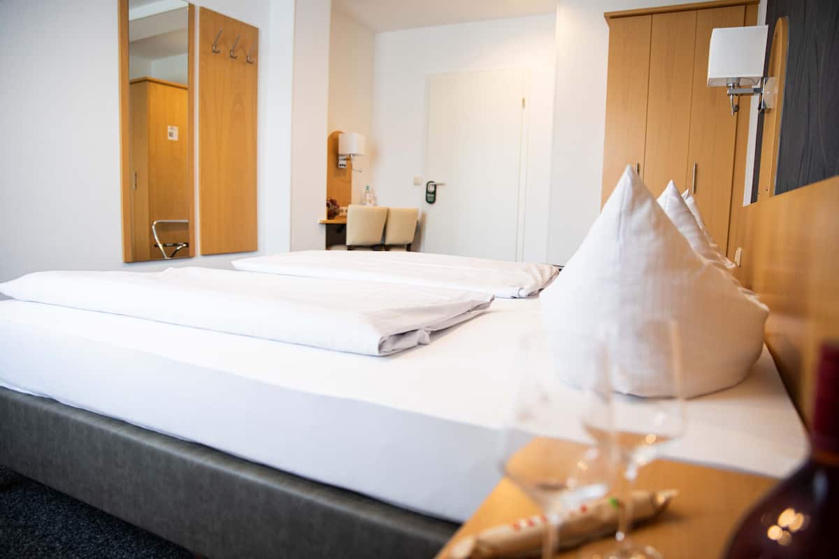 Quarto de hotel com duas camas, um guarda-roupa de madeira, uma escrivaninha com cadeira, um espelho e uma mesa de cabeceira com um copo de vinho.