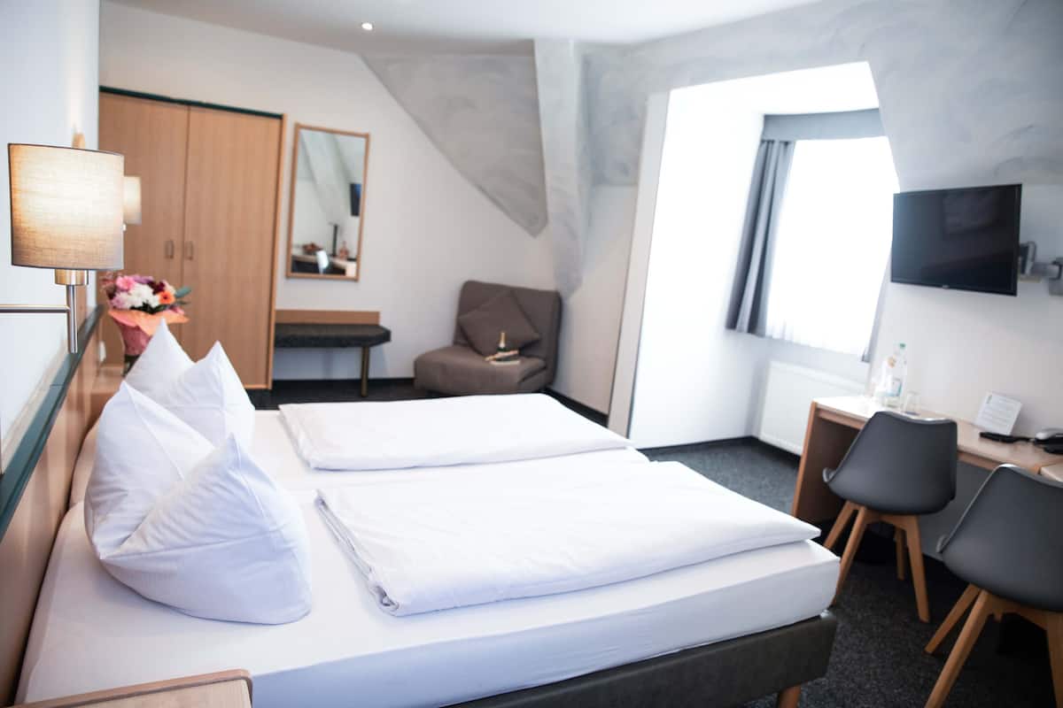 Quarto de hotel com cama, escrivaninha, cadeira, televisão, guarda-roupa e janela.