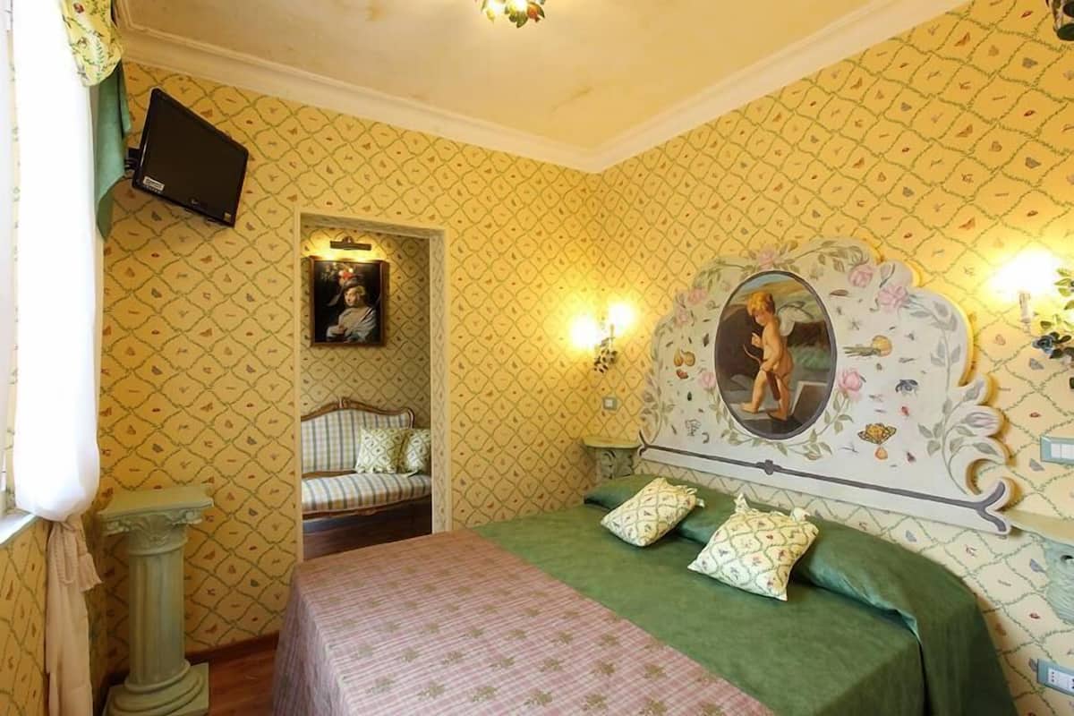 A bedroom with patterned wallpaper, a bed with green bedding, a mirror with a floral design, and a television mounted on the wall.