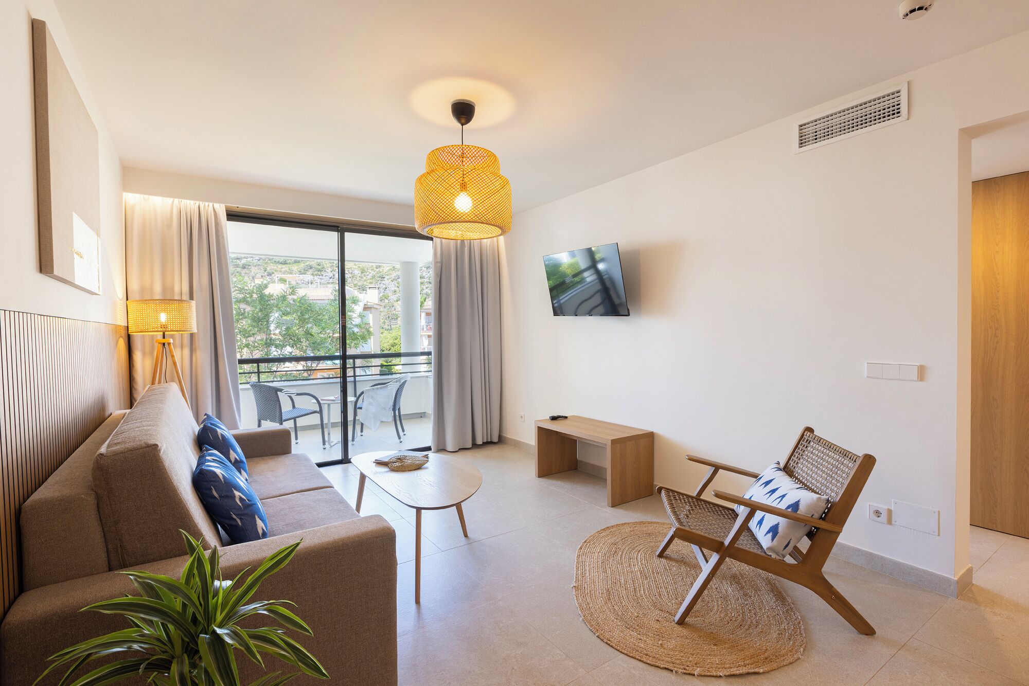 En moderne stue med sofa, sofabord, tv og en balkon med udsigt til grønne områder.