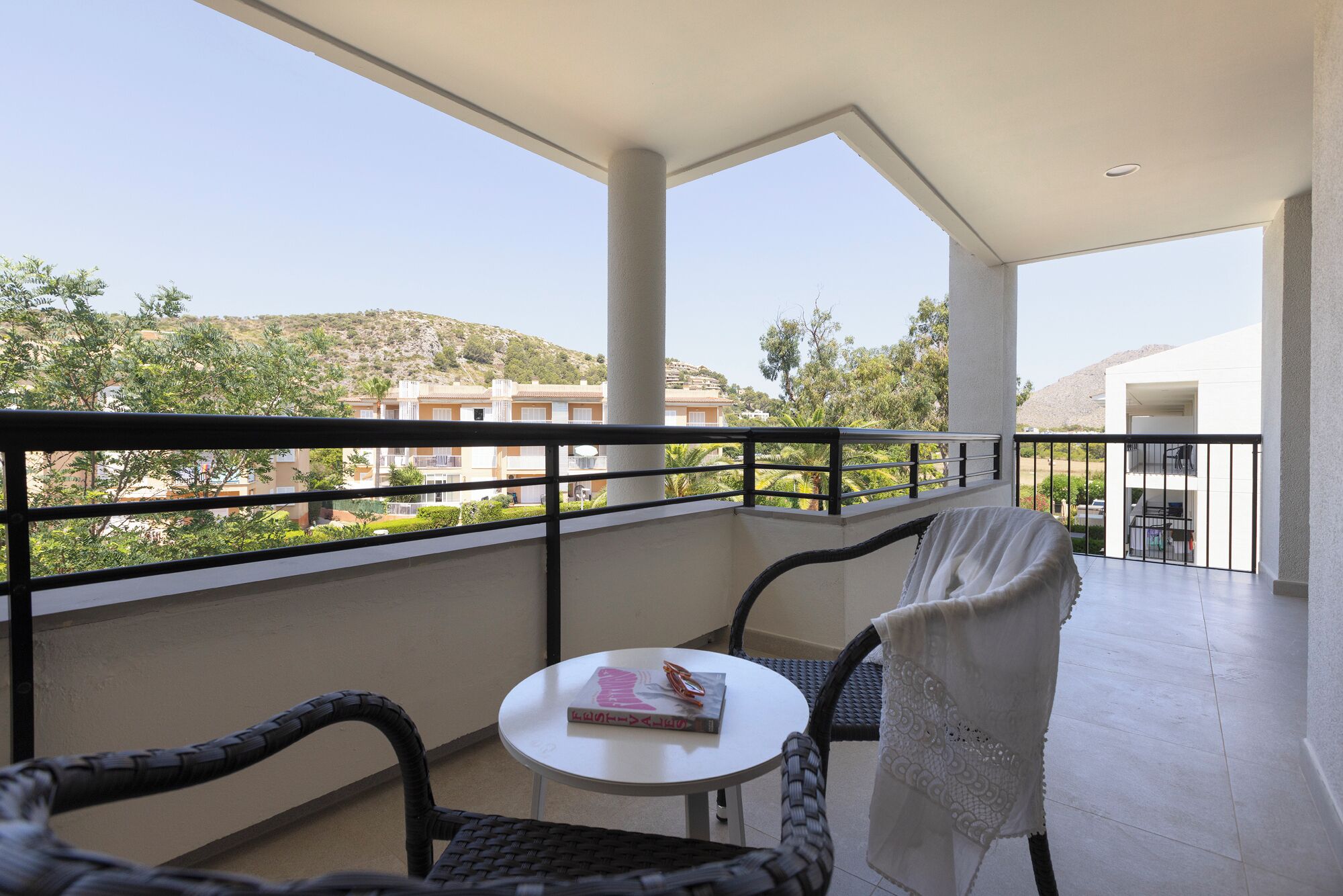 A balcony with wicker chairs, a small table, and a view of buildings and greenery.