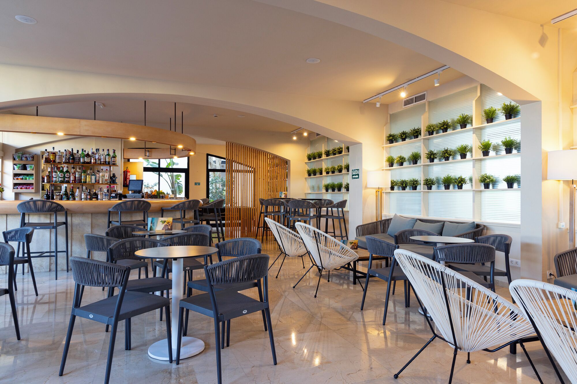 Zona bar moderna con soffitto curvo, sgabelli da bar e una varietà di bottiglie. Sono presenti aree salotto con tavoli e sedie, e una parete con mensole piene di piante in vaso.