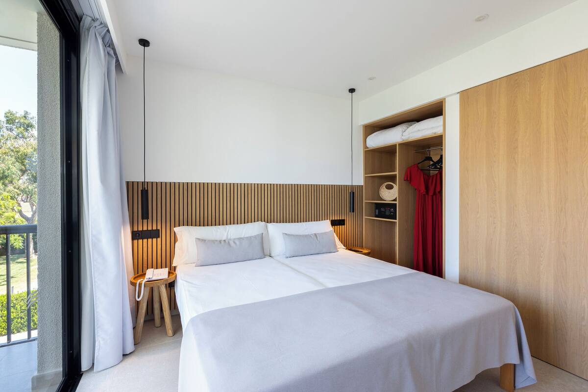 A modern hotel room with a large bed, a wooden headboard, a wardrobe, and a balcony.