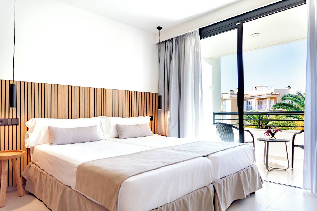 A modern hotel room with a large bed, a wooden headboard, a small table, and a balcony with a view of the city.