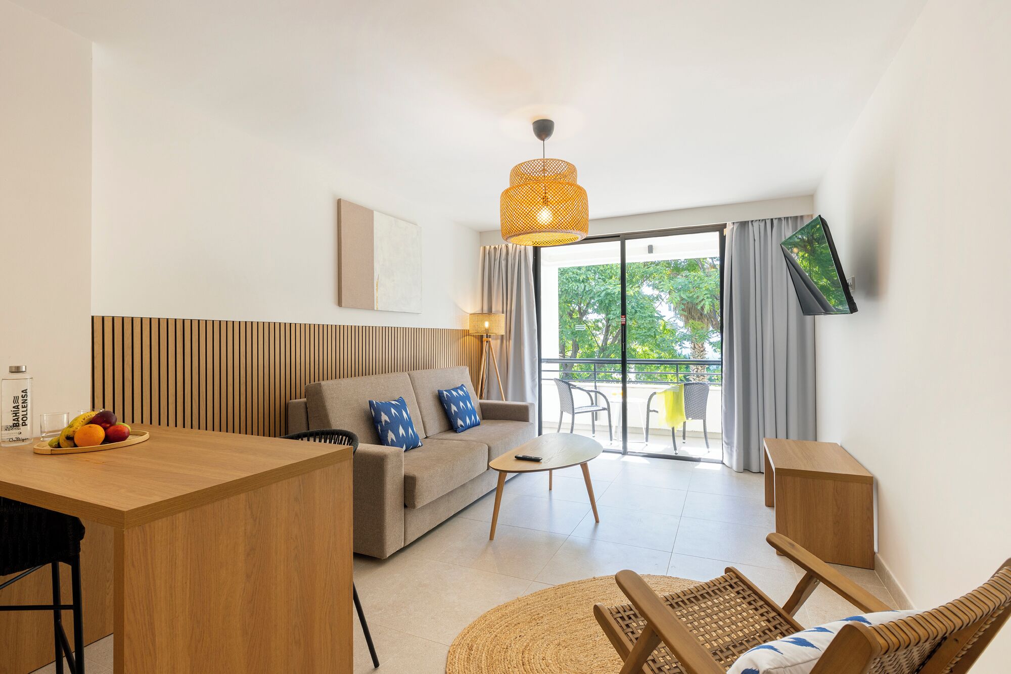 Una sala de estar moderna con una mesa de comedor de madera, un sofá con almohadones azules, una mesa de centro redonda y una silla de mimbre. Un televisor está montado en la pared, y se ve un balcón con vistas a árboles a través de puertas de vidrio.