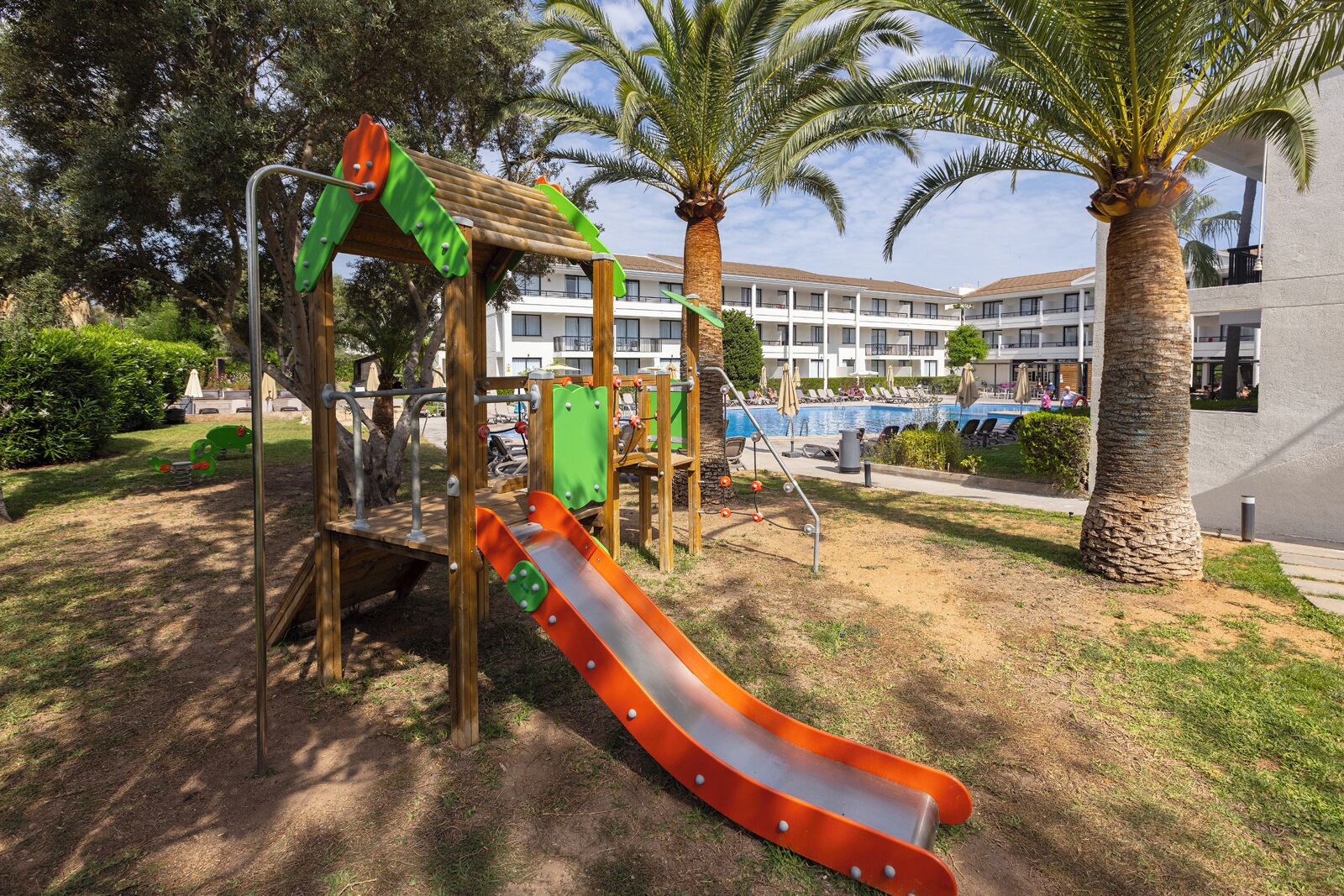 Un parque infantil con un tobogán y estructuras para escalar, rodeado de palmeras y una piscina al fondo.