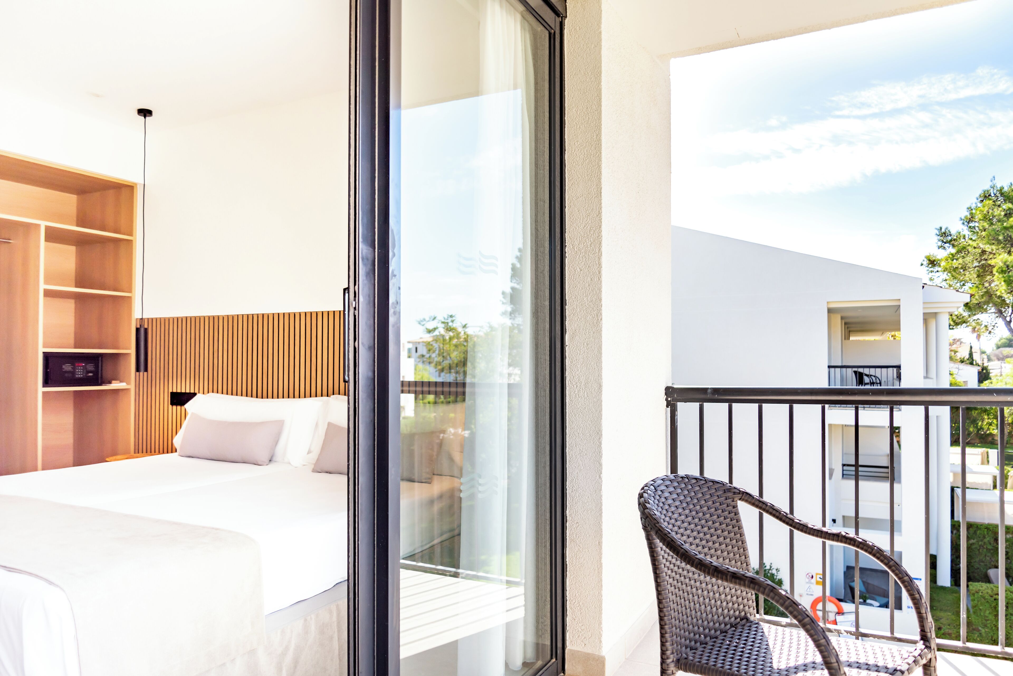 A hotel room with a bed, a wicker chair, and a balcony with a view of the outside.